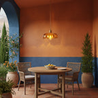 Amber Clérachic pendant glowing above a round wooden table on a terrace, against a terracotta and blue wall with potted plants.