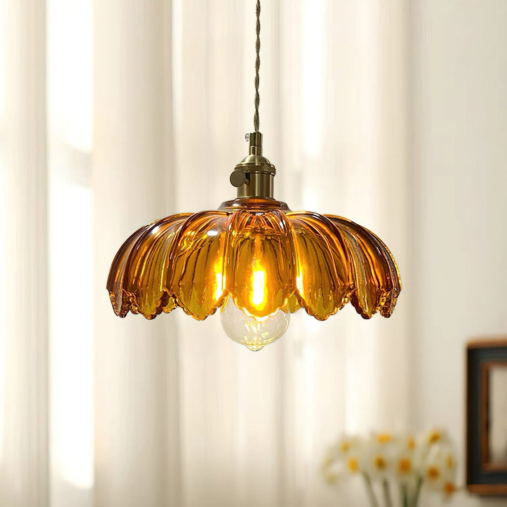 Close view of an amber glass Clérachic pendant with a ribbed, petal-edged shade, a brass switch fitting and a lit bulb.