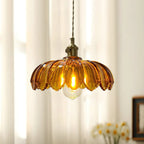 Close view of an amber glass Clérachic pendant with a ribbed, petal-edged shade, a brass switch fitting and a lit bulb.
