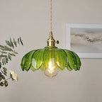 Lit green glass Clérachic pendant with a brass switch fitting, in front of a framed grey print and a sprig of eucalyptus.
