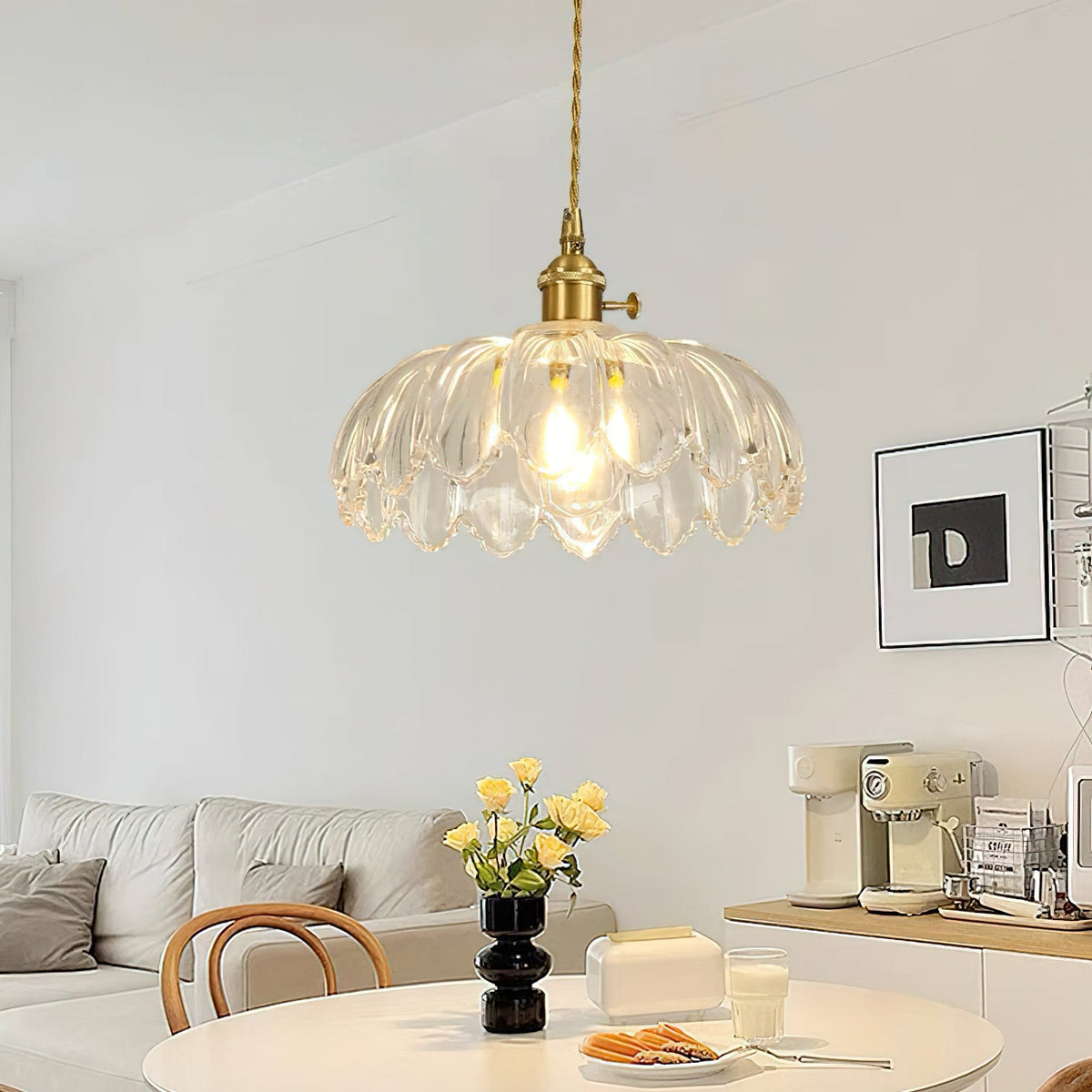 A clear glass Clérachic pendant with ribbed petals hangs above a round white table with flowers, a coffee machine behind.
