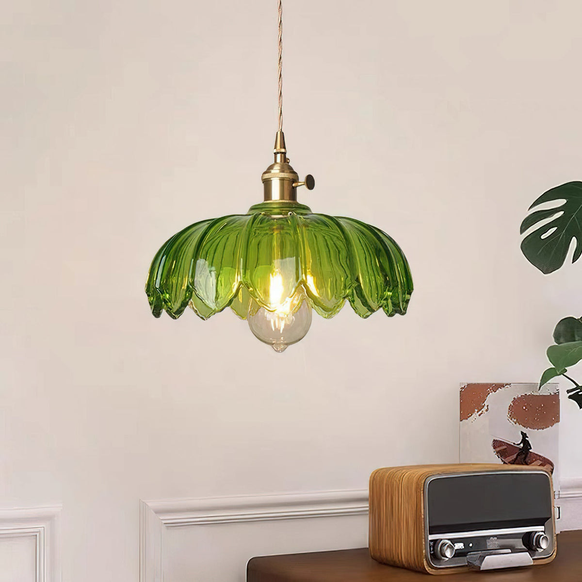 Green Clérachic pendant with a lit bulb, in front of a white panelled wall with a retro radio and a large leaf.