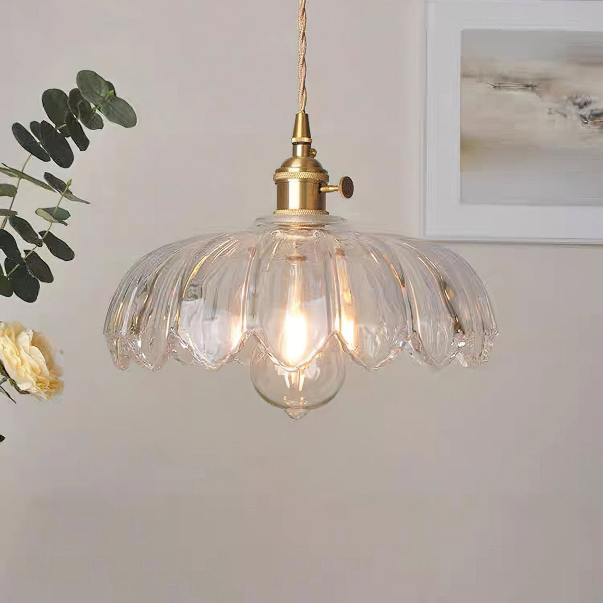 Clear glass Clérachic pendant with ribbed petals, lit by a filament bulb, with eucalyptus and a framed print behind.