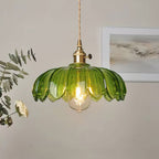 Lit green glass Clérachic pendant with a petal-edged shade and brass switch fitting, beside eucalyptus and a framed print.