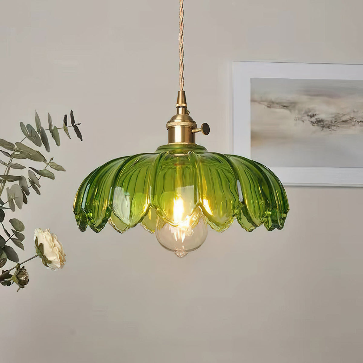 Lit green glass Clérachic pendant with a petal-edged shade and brass switch fitting, beside eucalyptus and a framed print.