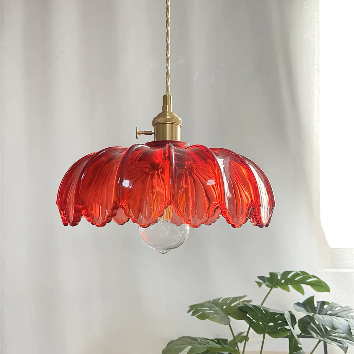 Red glass Clérachic pendant with ribbed petals and a brass switch, in front of a white curtain and a monstera leaf.