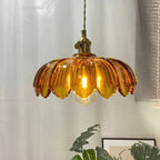 Amber Clérachic pendant glowing warm, with a bold petal-edged shade, in front of a sheer curtain and a green plant.