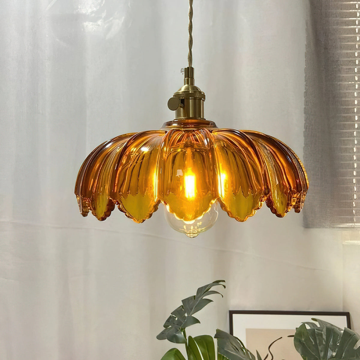 Amber Clérachic pendant glowing warm, with a bold petal-edged shade, in front of a sheer curtain and a green plant.