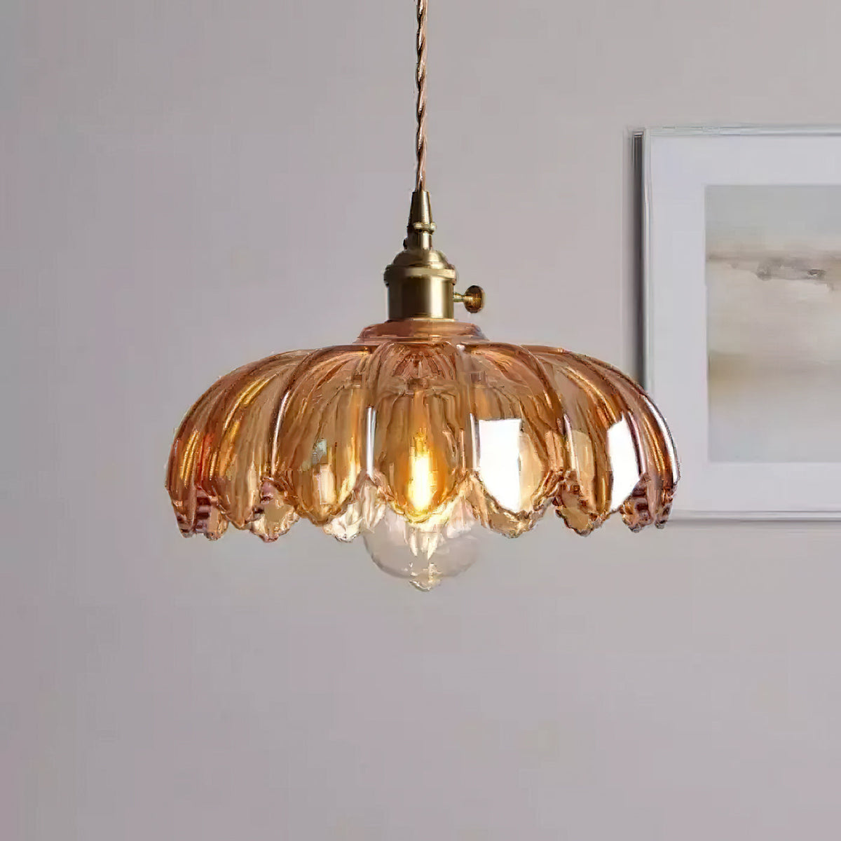 A pale amber-pink Clérachic pendant with a petal-edged shade and a lit bulb, in front of a framed grey print.