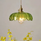 Green petal-edged glass Clérachic pendant with a lit bulb, above a spray of yellow flowers in a pale room.