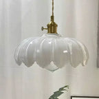 A white glass Clérachic pendant with a ribbed, petal-edged shade and a brass switch fitting, in front of a white curtain.