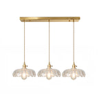 Row of three clear glass Clérachic pendants hanging from one long brass ceiling bar on a white backdrop.