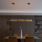 This Clérachic linear pendant glows over a dark wood dining room with a long table, tan chairs and a shelf of wine bottles.