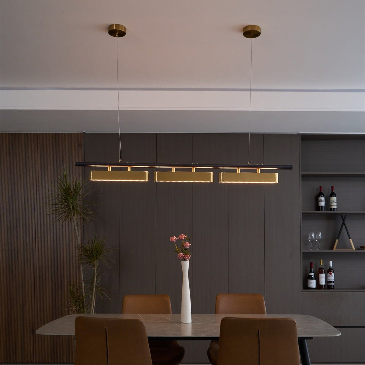 This Clérachic linear pendant glows over a dark wood dining room with a long table, tan chairs and a shelf of wine bottles.