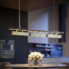 Clérachic linear pendant with three brass diffusers hung low over a dining table in a dark, moody kitchen with a round vase of white roses.