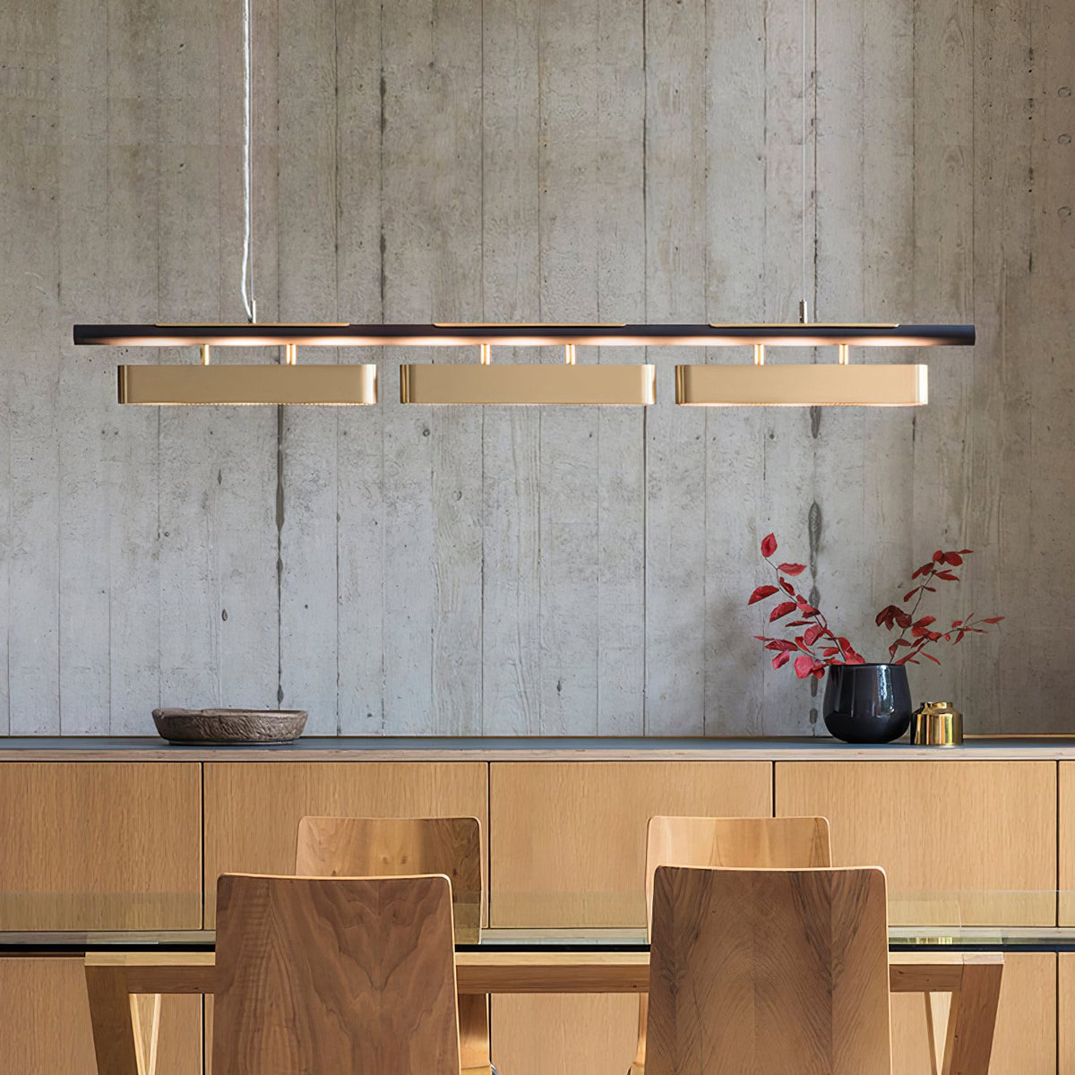 Clérachic brass and black linear pendant over a dining table, with a raw concrete wall and a sideboard of pale wood behind it.
