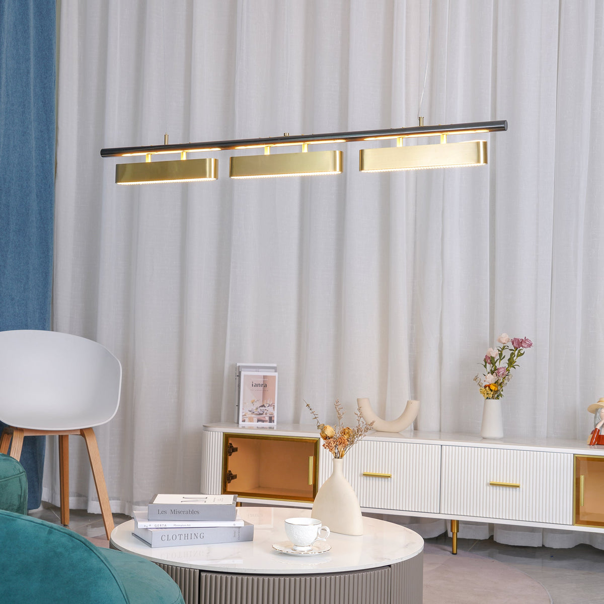 A Clérachic three-head linear pendant in front of sheer white curtains, above a living room with a white sideboard and a round table.