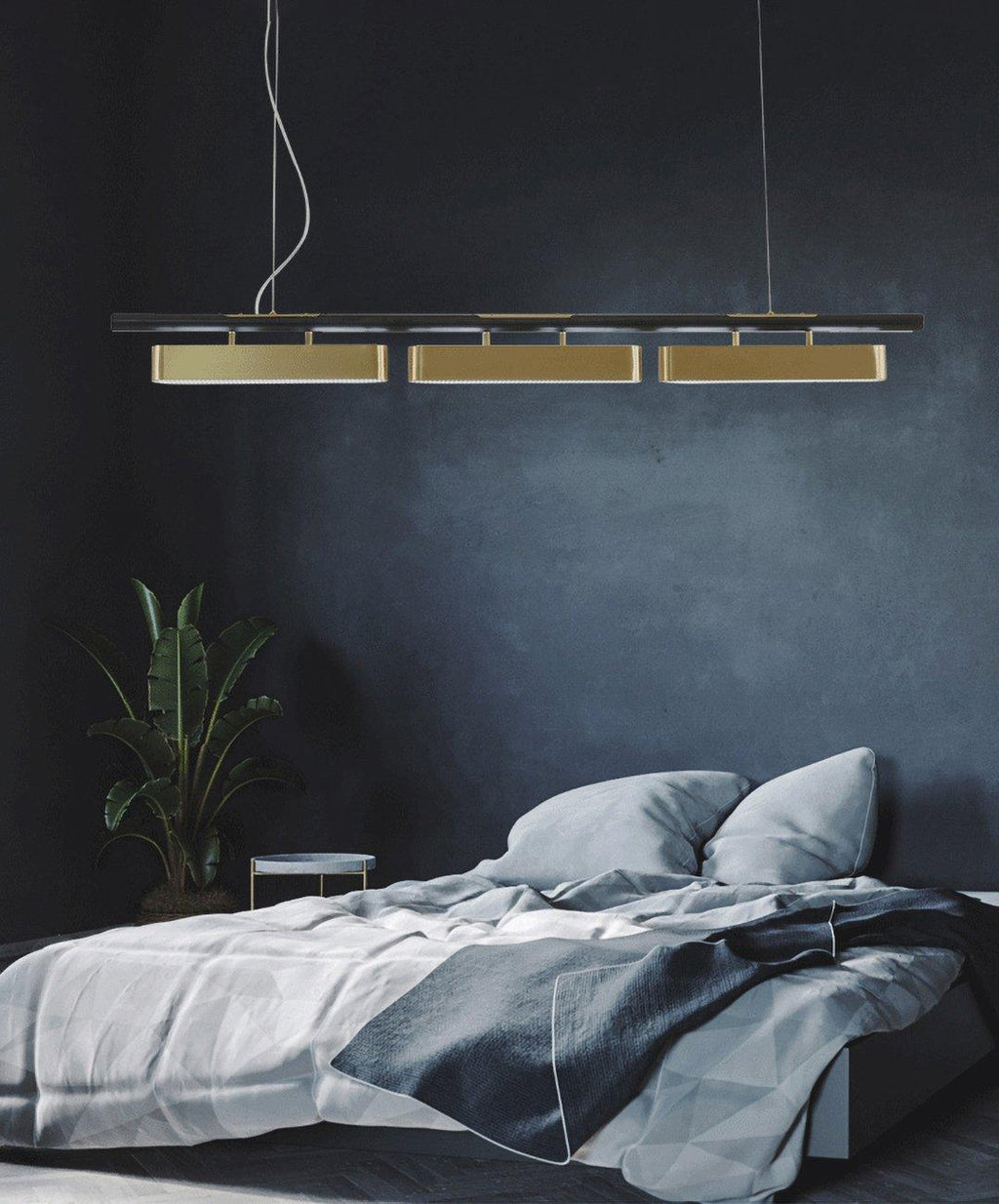A Clérachic three-head linear pendant hung against a dark blue wall above a bed with grey linen and a potted plant.