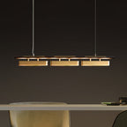 Clérachic linear pendant with three brass heads above a pale table, seen from the side against a dark grey wall.