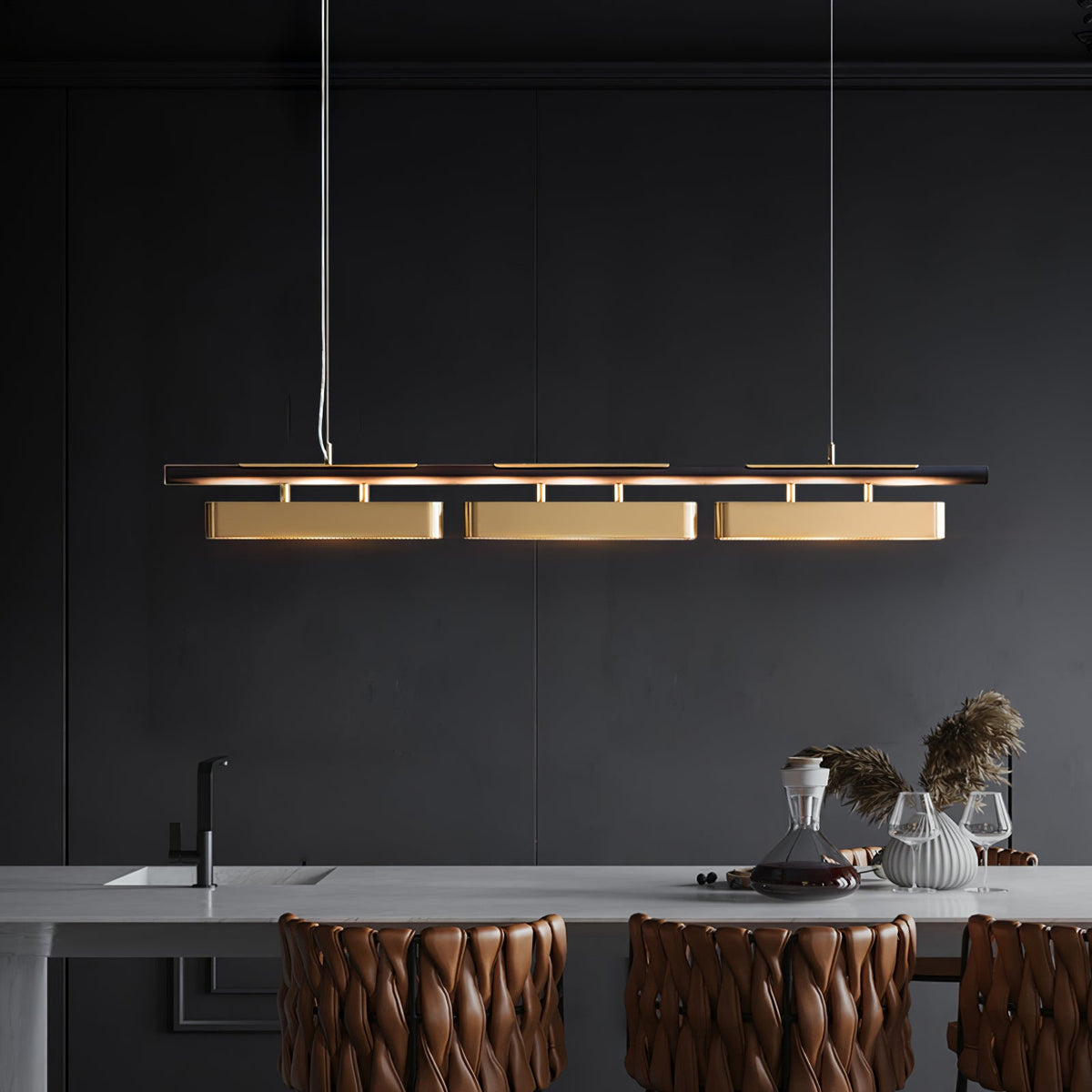 Three-head Clérachic brass pendant above a dark kitchen worktop with a decanter, dried grasses and woven leather stools.