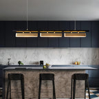 Wide view of the lit Clérachic three-head pendant over a kitchen island with black stools and a marble-effect splashback.