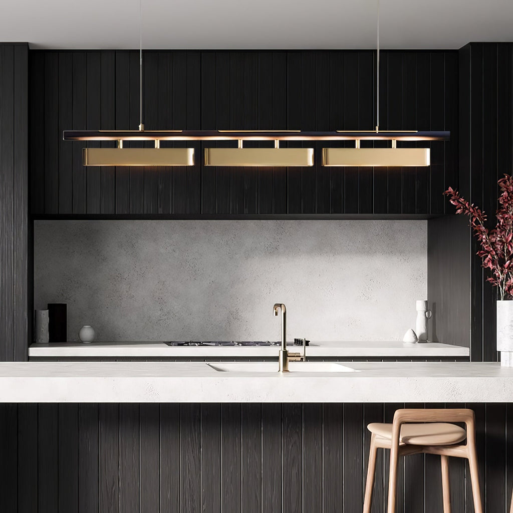 Clérachic linear pendant with three brass heads in a dark kitchen with a pale stone splashback, a brass tap and a wooden stool.