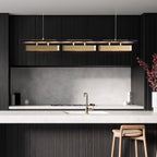 Clérachic linear pendant with three brass heads in a dark kitchen with a pale stone splashback, a brass tap and a wooden stool.