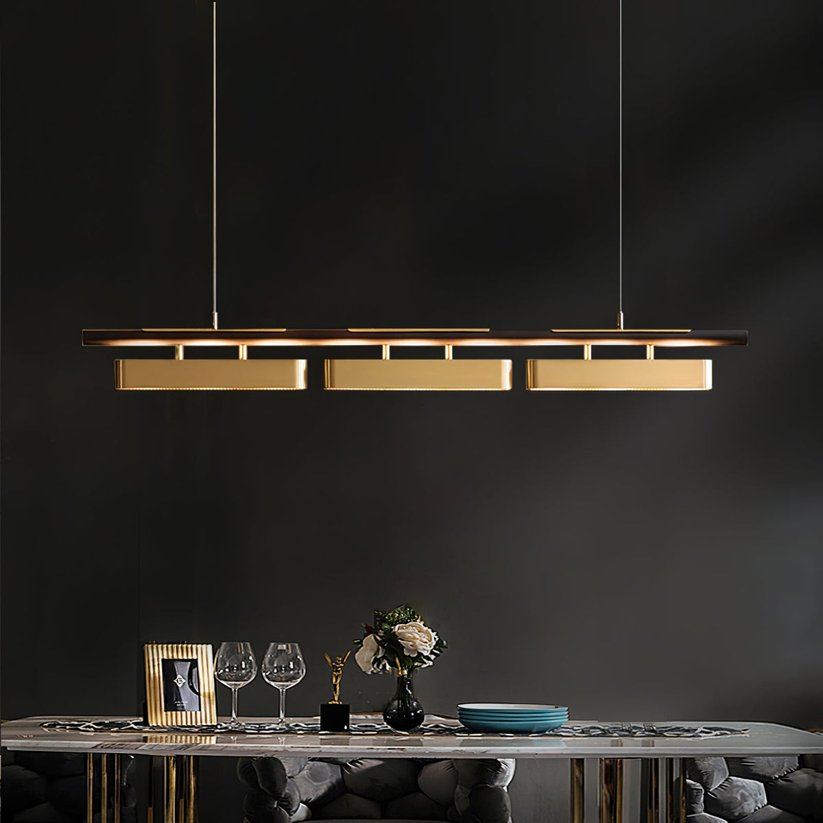 Clérachic brass pendant with three lit heads against a charcoal wall, above a console with glasses, flowers and a gold-framed photo.