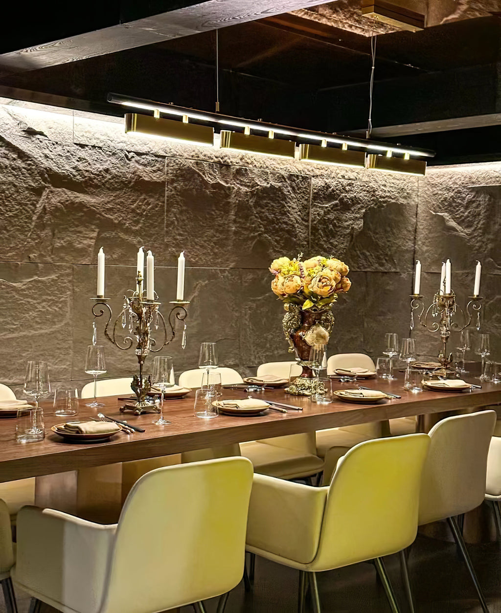 Clérachic brass pendant above a long dining table set with candles and flowers, against a rough stone wall and cream chairs.