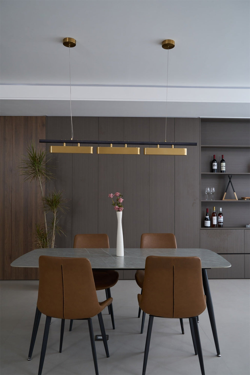 Clérachic three-head pendant in a dining room with a grey stone table, tan chairs, a white vase and a shelf of wine bottles.
