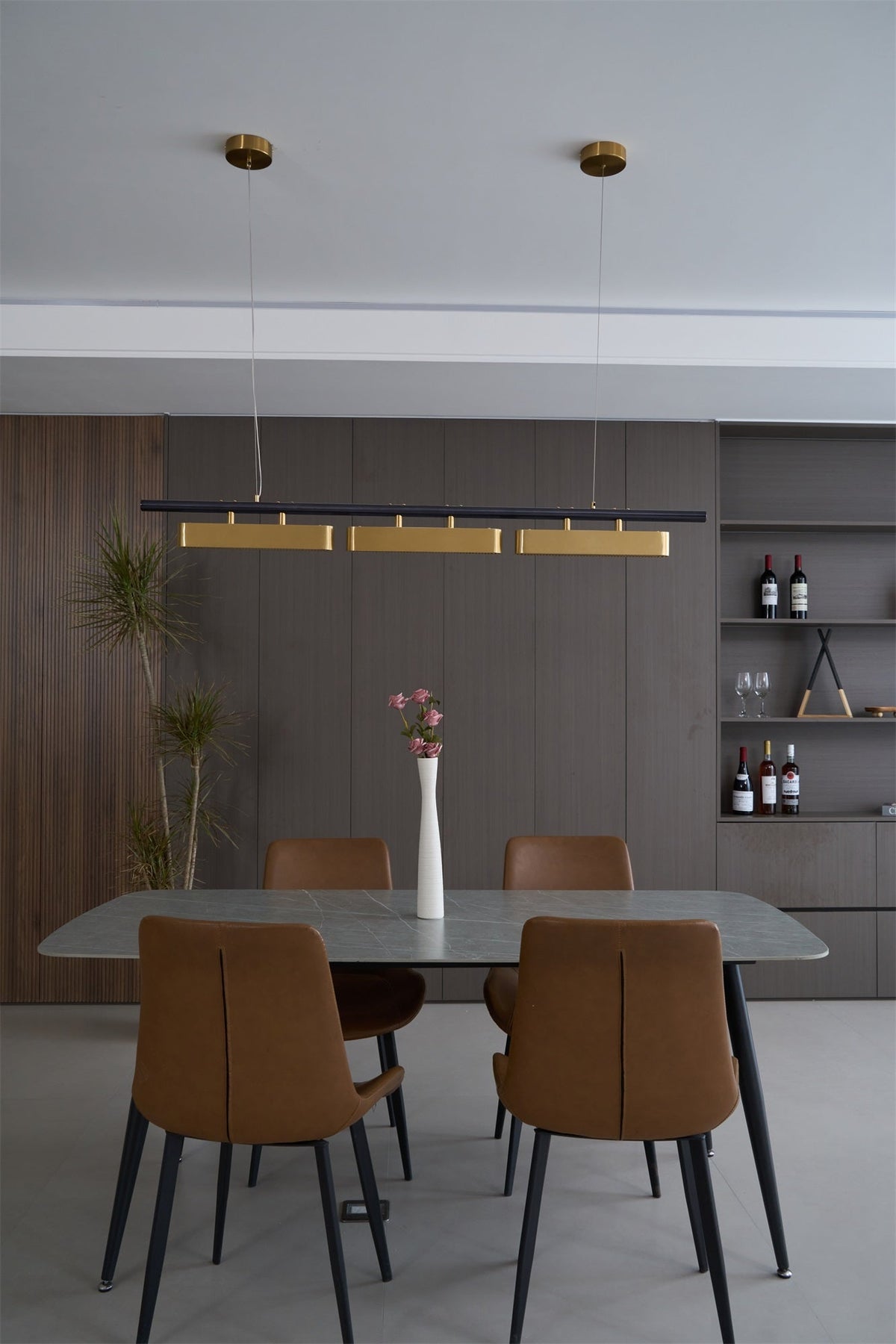 Clérachic three-head pendant in a dining room with a grey stone table, tan chairs, a white vase and a shelf of wine bottles.