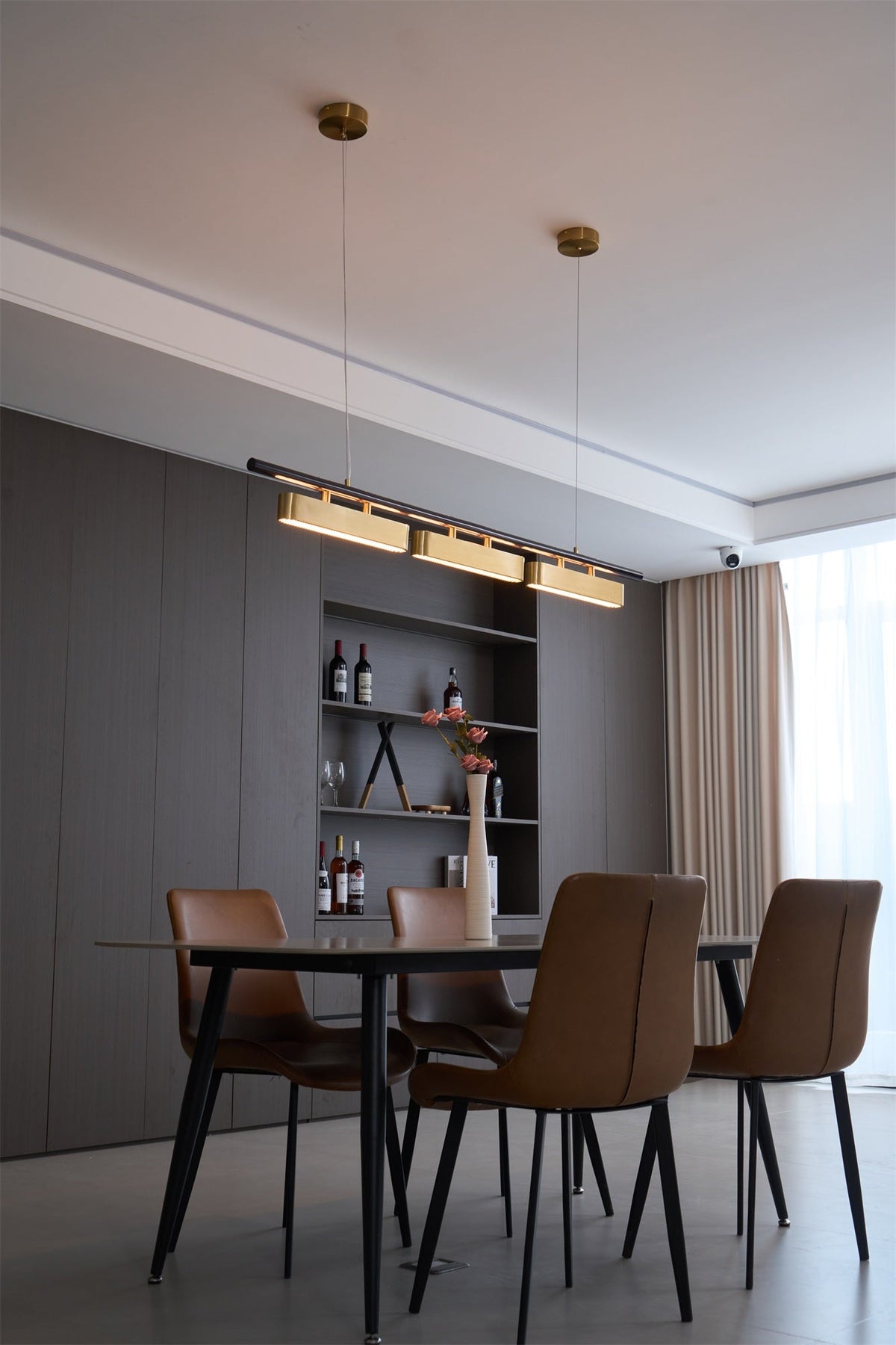 Distant view of a Clérachic pendant on two brass canopies, above a dining table with tan chairs and a wine shelf.