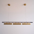Lit Clérachic three-head pendant on a pale wall, with a warm glow along the black bar and two round brass canopies above.