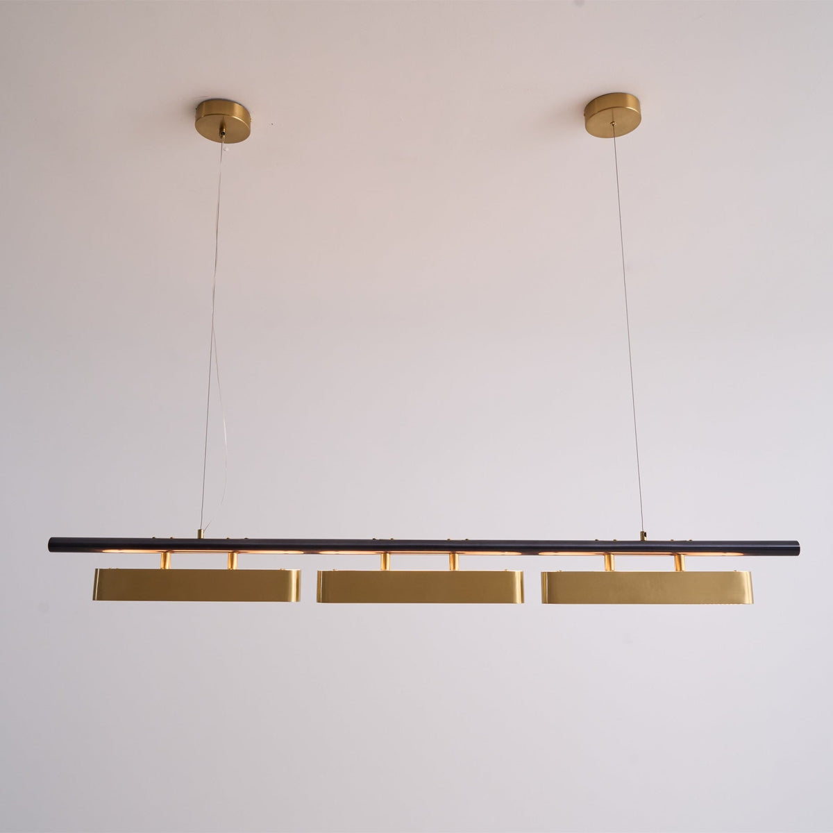 Lit Clérachic three-head pendant on a pale wall, with a warm glow along the black bar and two round brass canopies above.
