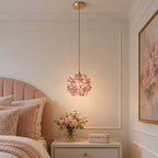 Lit pink glass petal Clérachic pendant hanging beside a blush headboard, above a white bedside table with a vase of roses.