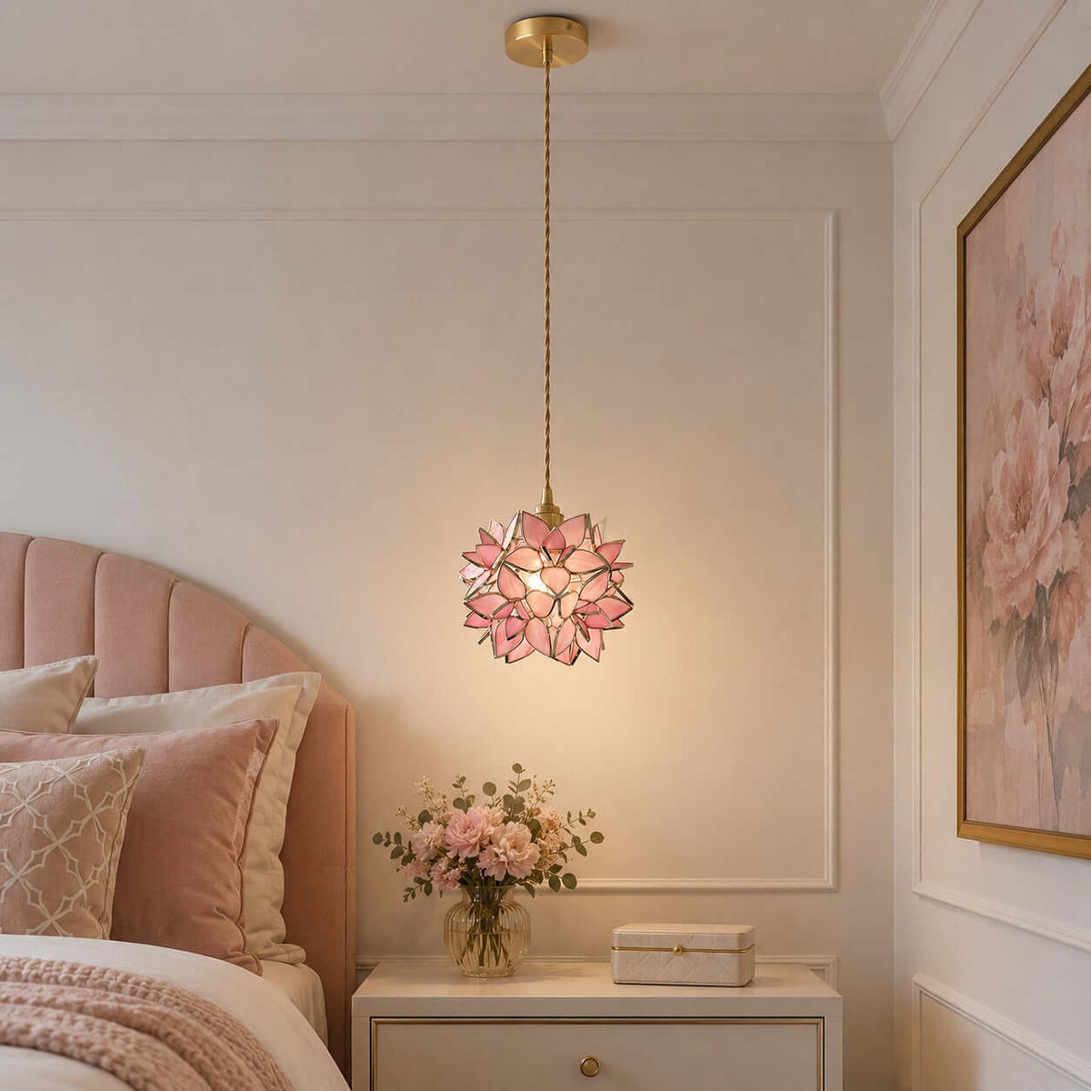 Lit pink glass petal Clérachic pendant hanging beside a blush headboard, above a white bedside table with a vase of roses.