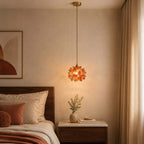 Clérachic pendant with an orange petal shade lit above a dark wood bedside table and a bed with terracotta cushions.