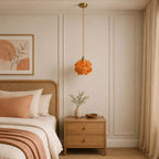 A small orange petal Clérachic pendant hangs on a brass fitting beside a cane bed, with a light oak bedside chest below.