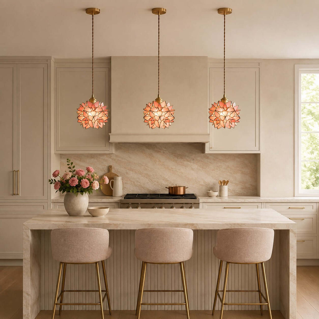 A row of three coral petal Clérachic pendants lit above a stone kitchen island and blush bar stools, with a copper pan behind.
