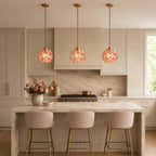 A row of three coral petal Clérachic pendants lit above a stone kitchen island and blush bar stools, with a copper pan behind.