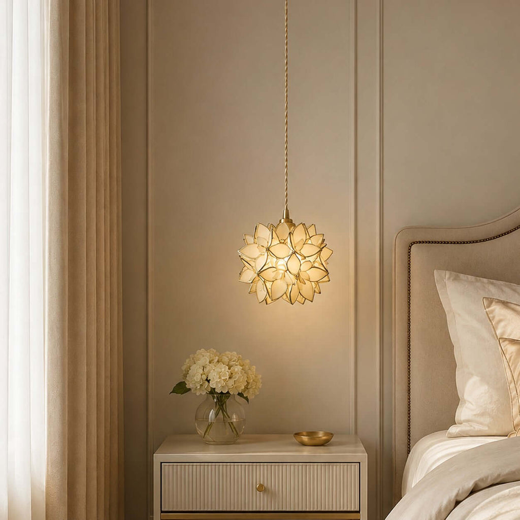 Warm light shines through the clear and gold-edged petals of this Clérachic pendant, beside a headboard and cream bedside table.