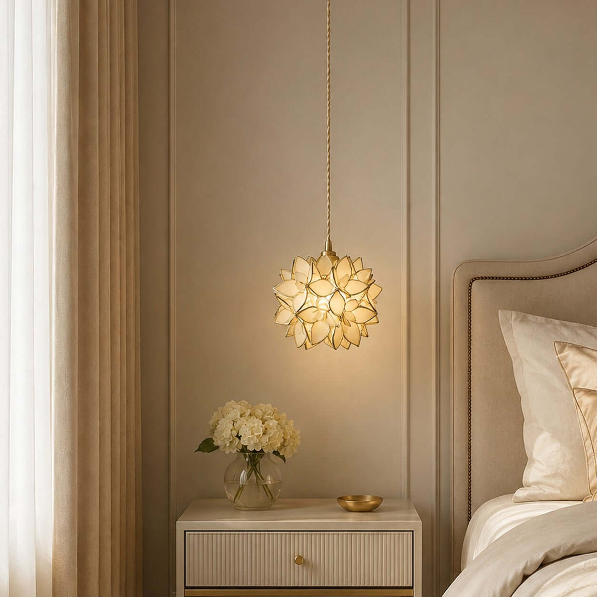 Warm light shines through the clear and gold-edged petals of this Clérachic pendant, beside a headboard and cream bedside table.