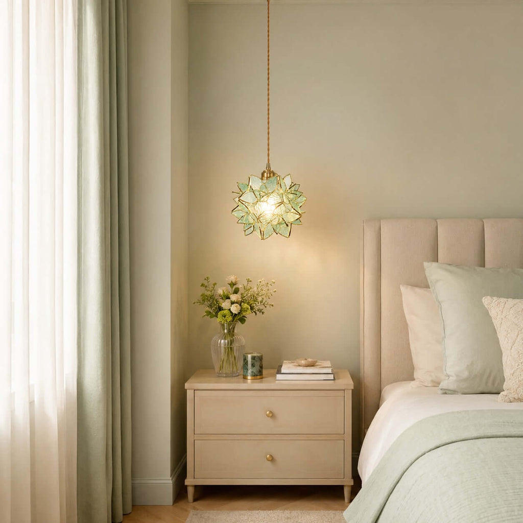 A pale green petal Clérachic pendant lit beside a bed with sage bedding, a cream two-drawer bedside table and sheer curtains.