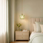 A pale green petal Clérachic pendant lit beside a bed with sage bedding, a cream two-drawer bedside table and sheer curtains.