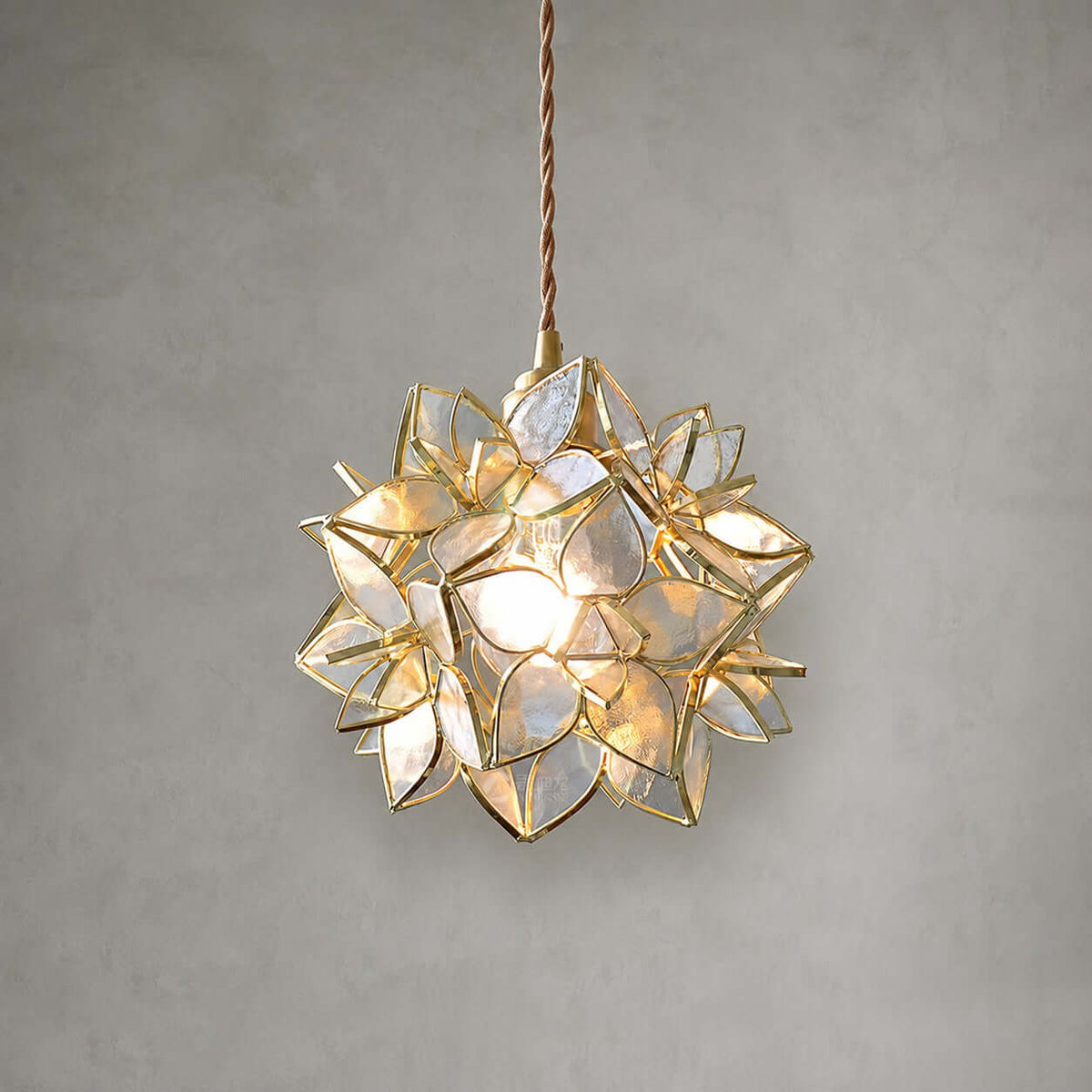 Clear glass petals with gold edging form this lit Clérachic pendant, which hangs on a braided cord against a grey wall.