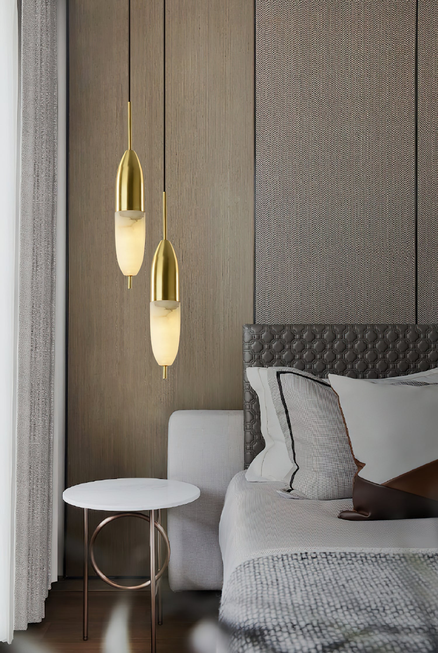 Pair of Clérachic Brass Pendant Lights over a round marble table with a brass ring base, beside a tufted grey bed.