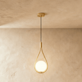 Clérachic brass teardrop wire pendant holding a glowing opal glass globe, hung on a black cord from a round brass ceiling plate.