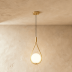 Clérachic brass teardrop wire pendant holding a glowing opal glass globe, hung on a black cord from a round brass ceiling plate.