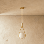 Clérachic teardrop pendant in brushed brass with an opal white globe, switched off, against a soft plaster wall and ceiling.
