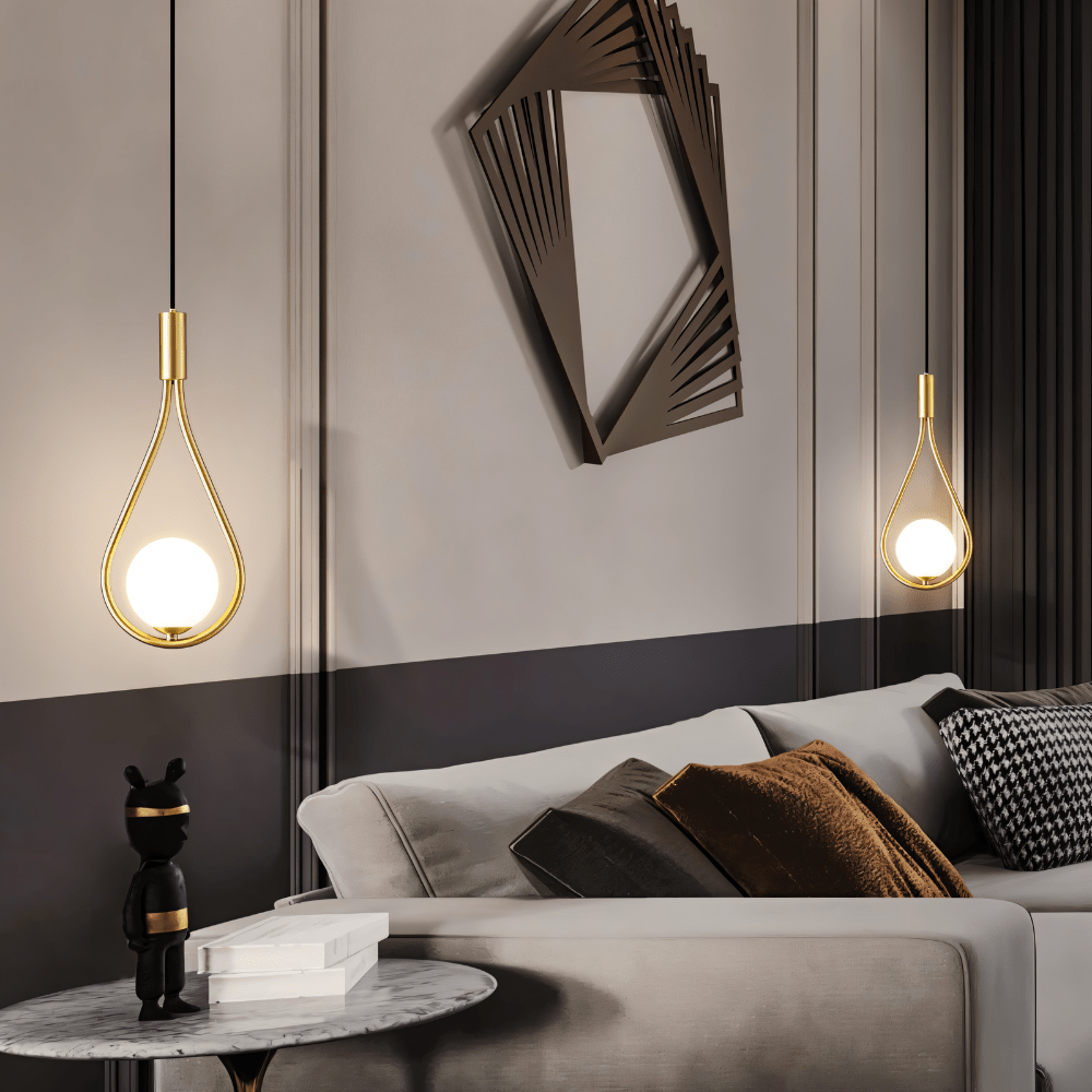Two Clérachic brass teardrop pendants with glowing opal globes hung at different heights beside a grey sofa and marble side table.